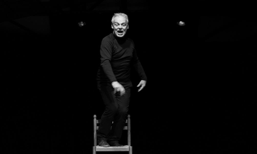 Nicolas Rassat debout sur une chaise, seul en scène dans la pénombre